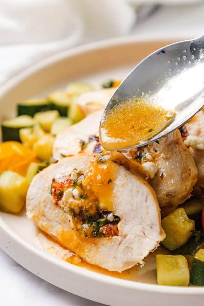A close-up of Mediterranean Stuffed Chicken breast slices on a plate with mixed vegetables, as a spoon drizzles golden sauce over the top. The flavorful stuffing includes Mediterranean herbs and cheese.