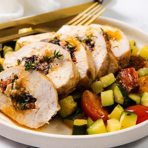 Mediterranean Stuffed Chicken breast, sliced and garnished with herbs, is served alongside chopped zucchini, yellow squash, and cherry tomatoes. A gold fork and knife rest at the top of the plate.