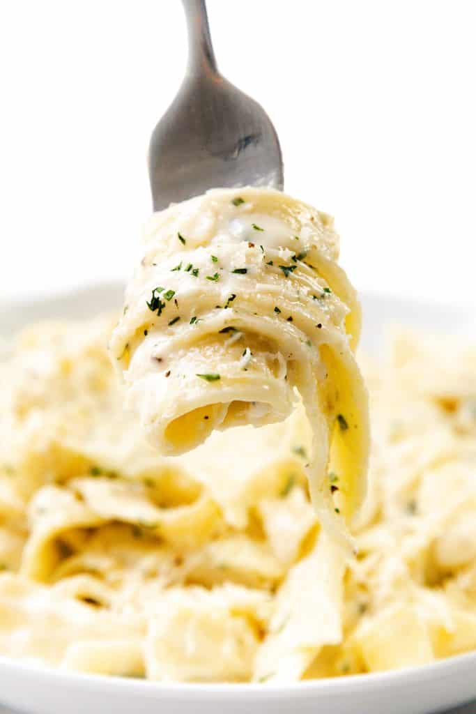 A fork lifts a swirl of creamy garlic fettuccine Alfredo, garnished with chopped parsley, with more pasta blurred in the background on a white plate—deliciously simple as a one pot meal.