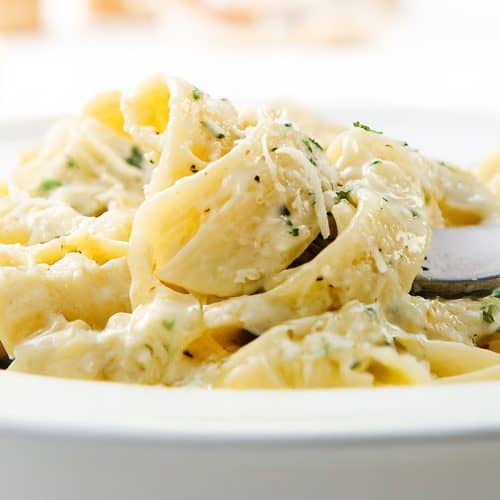 Close-up of creamy garlic fettuccine Alfredo pasta topped with grated cheese and herbs in a white bowl, with a fork twirling some noodles.