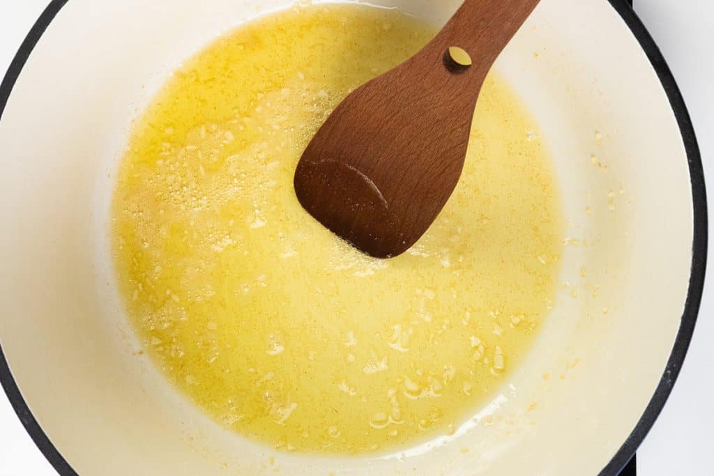 A wooden spoon stirs minced garlic cooking in melted butter inside a white enameled pot, the first step to a rich and creamy One Pot Alfredo.