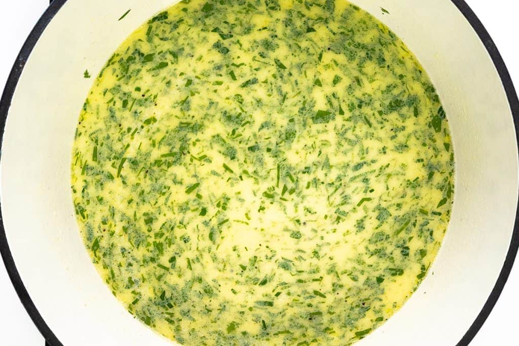 A close-up top view of a pot filled with creamy garlic fettuccine alfredo soup, featuring finely chopped green herbs that create a speckled yellow and green appearance.