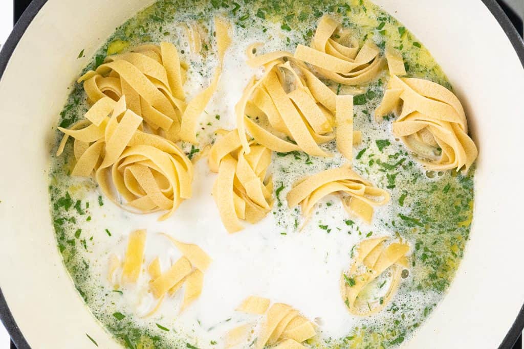 A pot filled with uncooked tagliatelle nests, creamy broth, and chopped green herbs, ready to become a delicious one pot fettuccine alfredo.