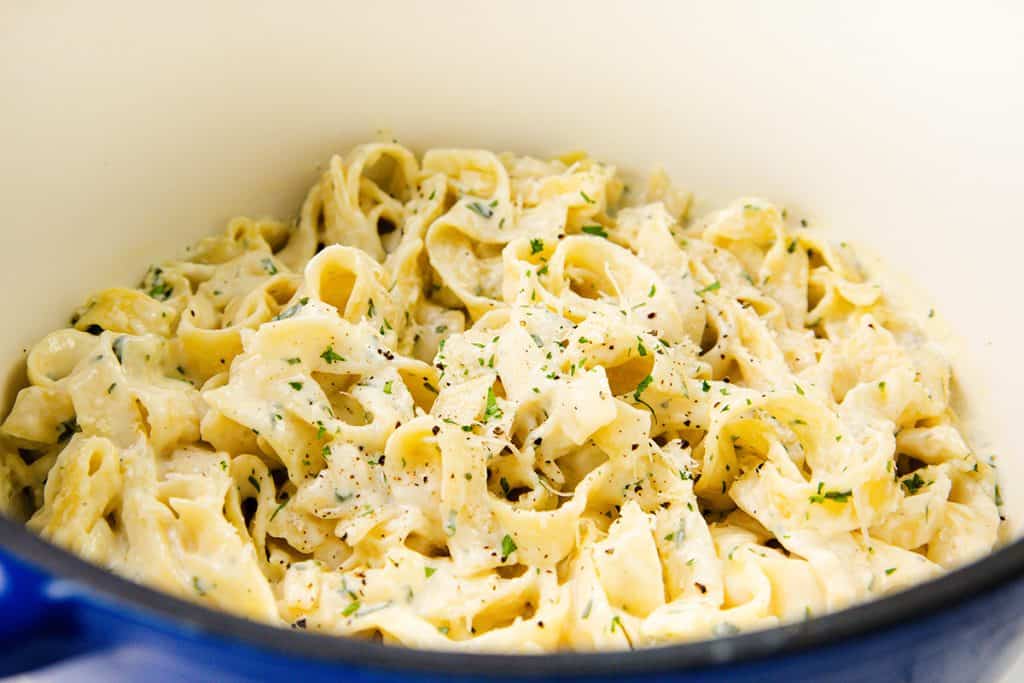 A close-up of creamy garlic fettuccine Alfredo pasta, garnished with chopped parsley and cracked black pepper, served in a white one pot.