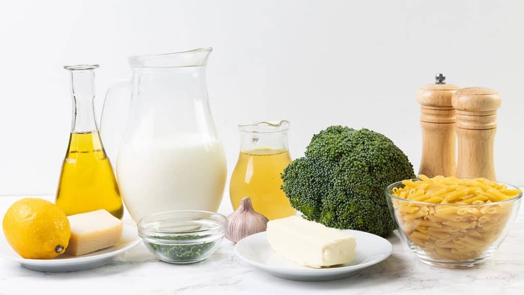 A variety of ingredients for a creamy garlic pasta on a white surface, including lemon, cheese, olive oil, milk, herbs in a bowl, garlic, butter, broccoli, penne pasta, another liquid in a small jug, and salt and pepper shakers.