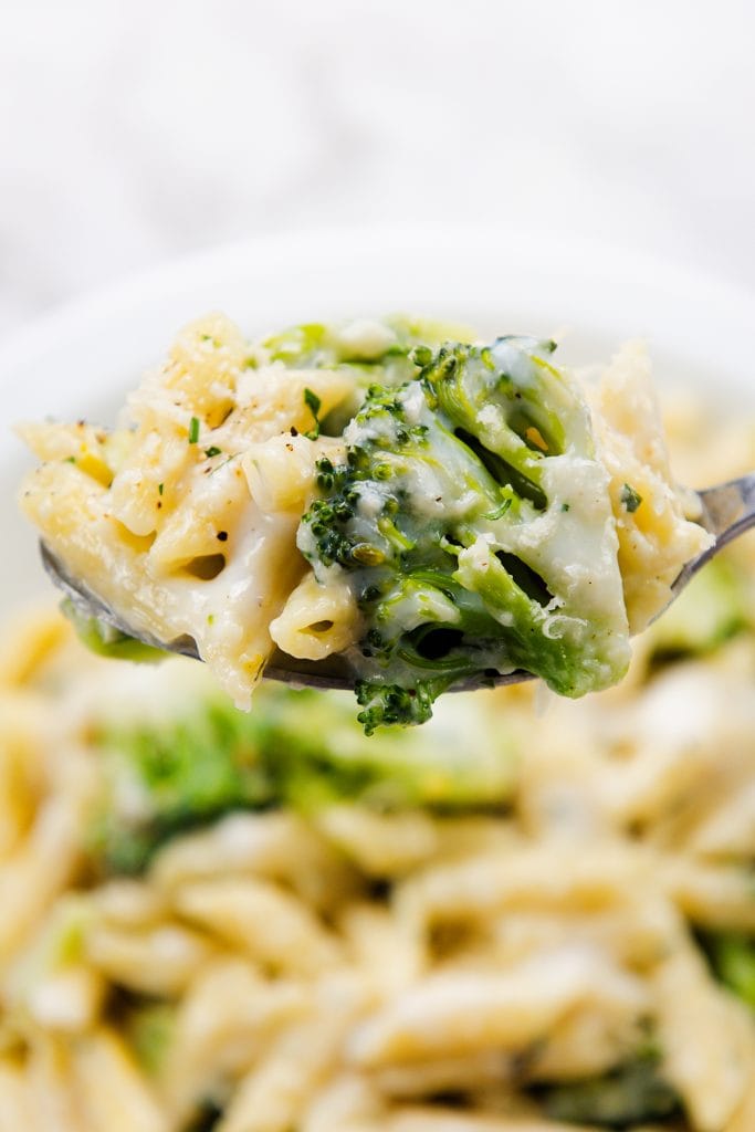 A close-up of a fork holding creamy garlic pasta penne with broccoli, covered in a white cheese sauce, with more pasta and broccoli visible in the blurred background.
