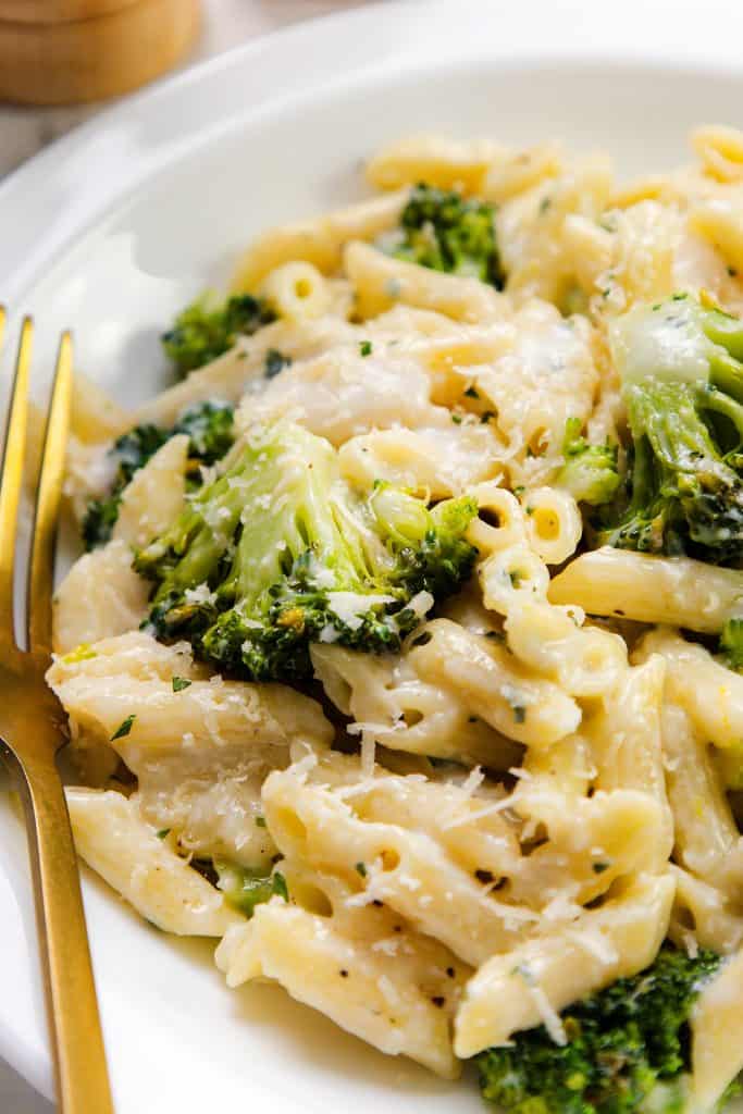 A close-up of One-Pot Pasta with penne and broccoli florets, all coated in a creamy garlic sauce, served on a white plate with a gold fork beside it.