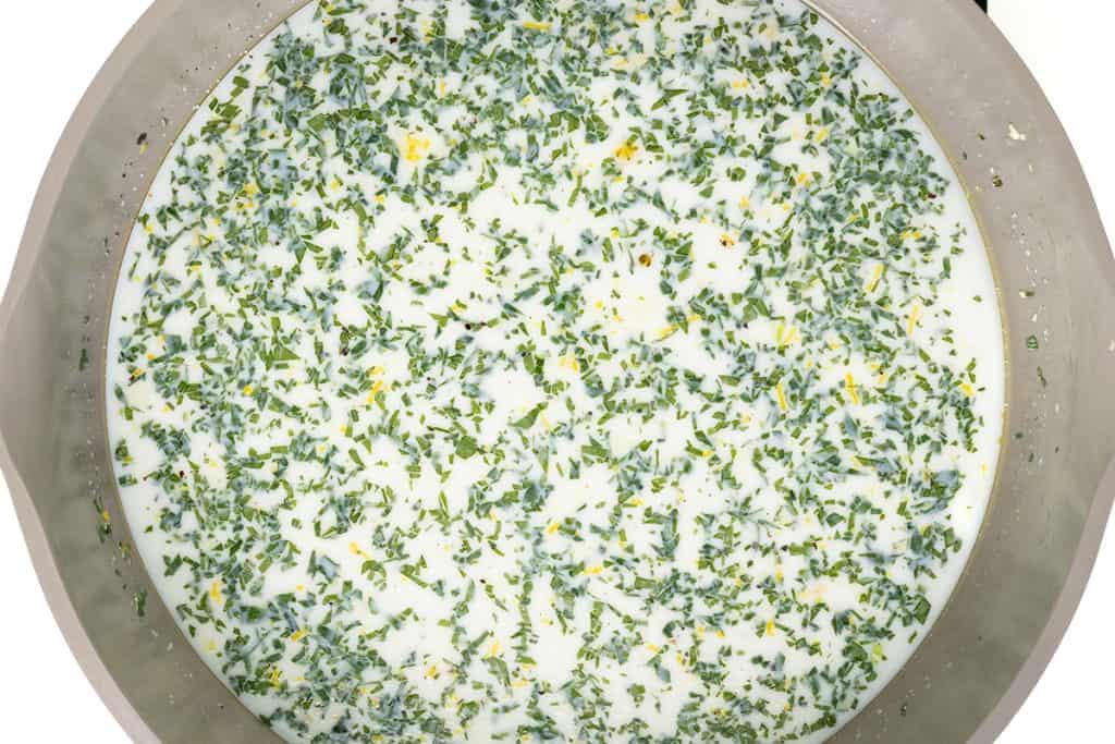 A close-up of a pot filled with creamy garlic pasta, its velvety white sauce speckled with finely chopped green herbs and yellow zest, makes this one-pot dish irresistibly inviting.