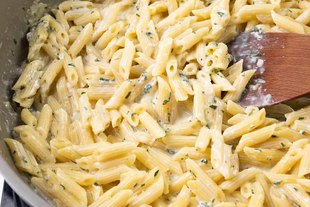 Close-up of creamy garlic pasta and penne mixed with chopped herbs, being stirred with a wooden spatula in a pan. This rich one-pot pasta features a thick sauce that coats every piece evenly.