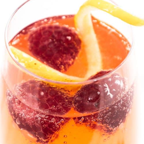 A close-up of a sparkling Raspberry Cocktail in a glass with fresh raspberries and a twist of lemon peel. Bubbles are visible on the surface, giving this Prosecco Cocktail a refreshing appearance.