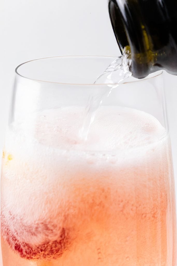 Close-up of a sparkling Raspberry Cocktail being poured from a bottle into a glass, with bubbles forming on top and a red fruit visible at the bottom of the light pink beverage.