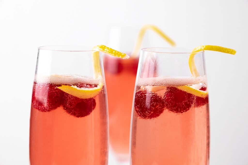 Three clear glasses filled with a pink Raspberry Blush sparkling drink, each garnished with raspberries and a curled lemon peel on the rim, against a white background.