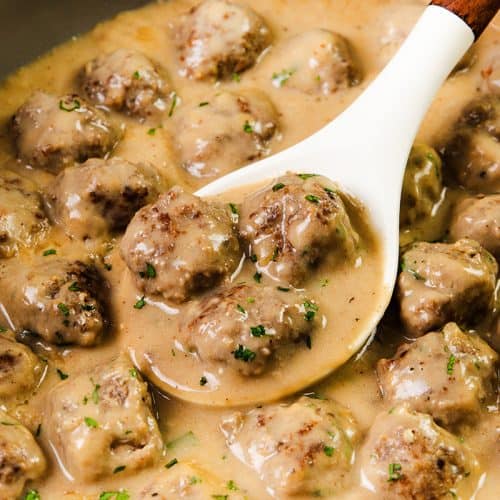 A white spoon scoops up creamy, saucy Swedish Meatballs garnished with chopped herbs from a pot filled with more meatballs in a rich, creamy gravy.
