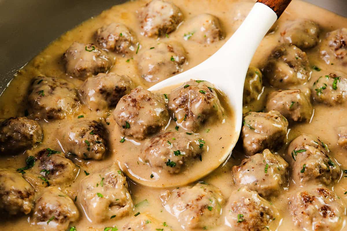 A white spoon scoops up creamy, saucy Swedish Meatballs garnished with chopped herbs from a pot filled with more meatballs in a rich, creamy gravy.