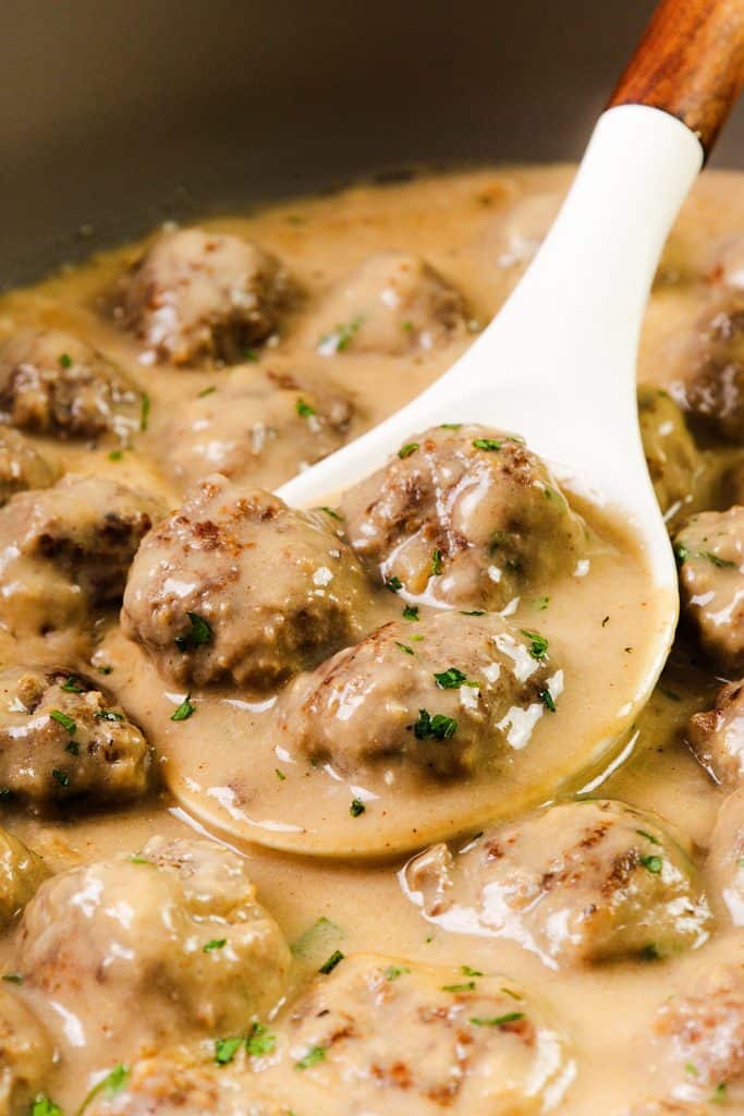A white spoon lifts Swedish Meatballs covered in creamy brown gravy, garnished with chopped parsley, from a pot filled with more meatballs and sauce.