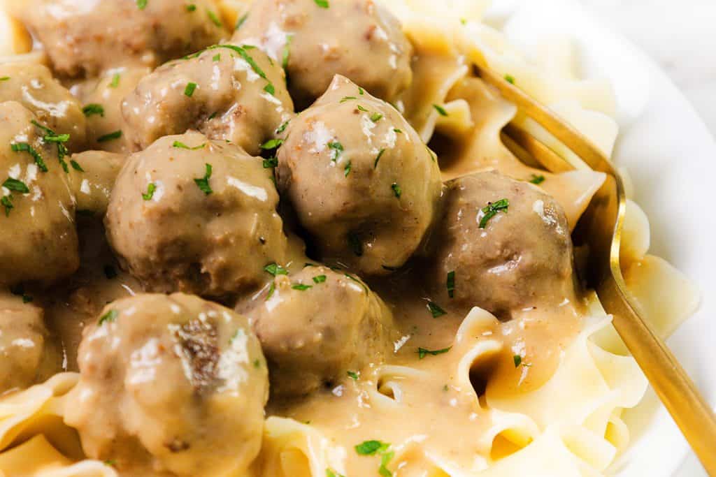 A close-up of Swedish Meatballs covered in creamy gravy, served over wide egg noodles, garnished with chopped parsley, with a gold fork on the side.