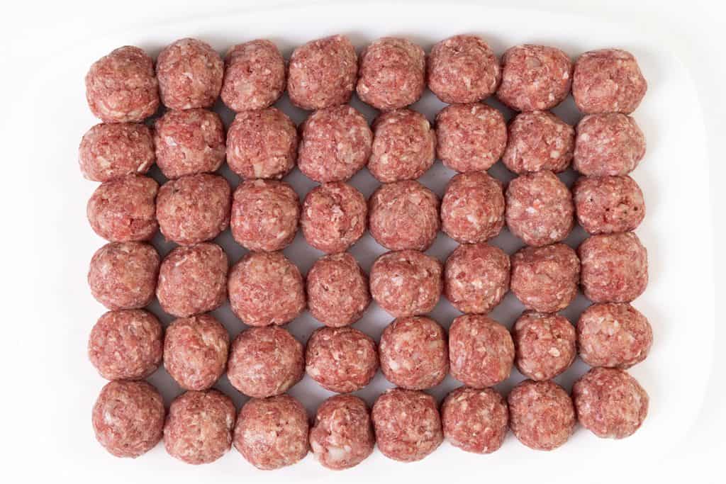 A white tray filled with neatly arranged rows of uncooked Swedish Meatballs made from ground meat. The uniform meatballs are ready to be cooked and served with a classic creamy gravy, filling the tray in a perfect grid pattern.