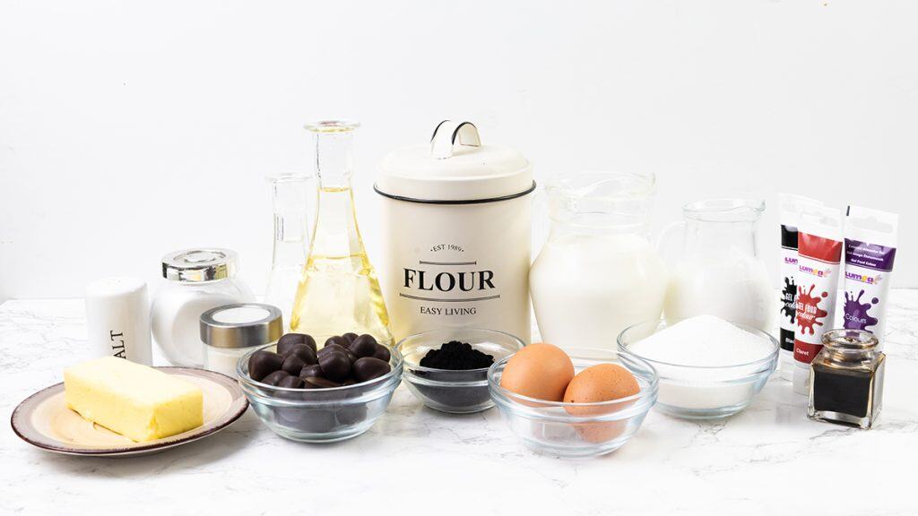 A variety of baking ingredients on a white surface, perfect for making Black Valentine&rsquo;s Day Cupcakes, including flour, eggs, butter, sugar, salt, oil, chocolate, milk, cream, black food coloring, vanilla, and red and purple gel food coloring.