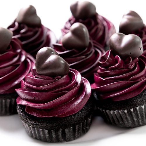 Six Black Valentine&rsquo;s Day Cupcakes with dark purple frosting and chocolate heart decorations are arranged closely together on a white plate.