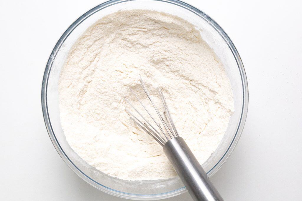 A glass bowl filled with flour and a metal whisk resting inside, all on a white surface&mdash;perfect for mixing up batter for Black Valentine&rsquo;s Day Cupcakes.