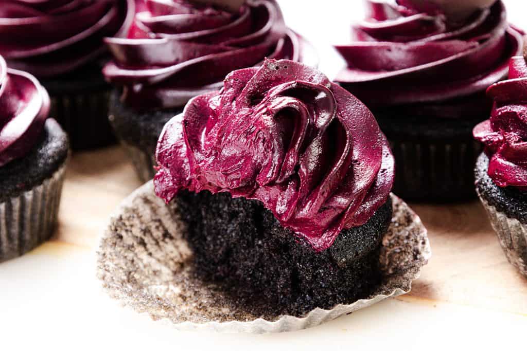 A close-up of Black Valentine&rsquo;s Day Cupcakes with dark purple frosting. One cupcake, partially unwrapped, has a bite taken out, revealing a moist, rich chocolate interior. The frosting is swirled decoratively on top.