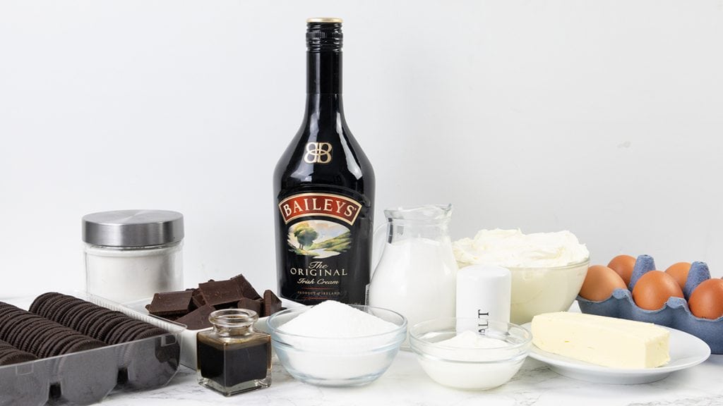 A variety of baking ingredients are arranged on a white surface, including a bottle of Baileys Irish Cream, eggs, butter, chocolate, and more&mdash;perfect for crafting a rich Irish Cream Cheesecake or other decadent treats.