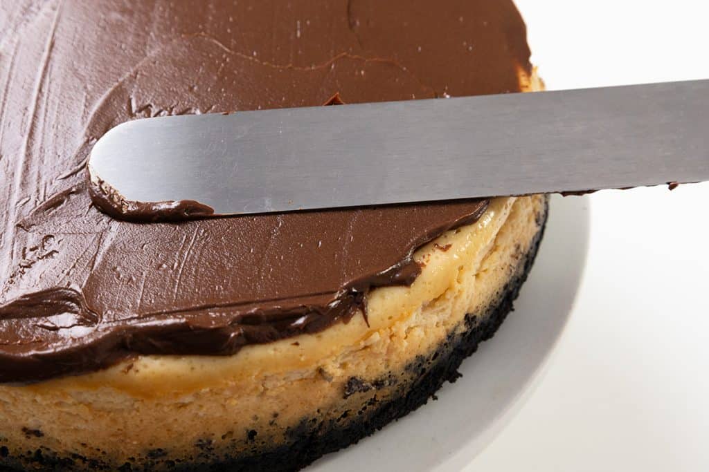 A metal spatula spreads smooth chocolate frosting over the top of an Irish Cream cheesecake with an Oreo cookie crust, sitting on a white plate.