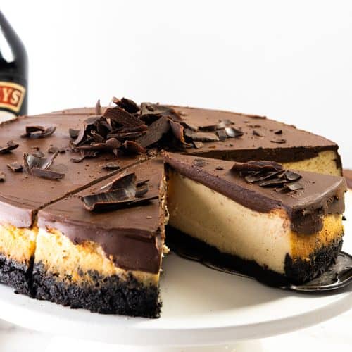 A chocolate-topped Irish Cream cheesecake with an Oreo crust sits on a white cake stand. A slice is being served, revealing creamy layers. Chocolate curls decorate the top, and a bottle of Baileys Irish Cream is in the background.