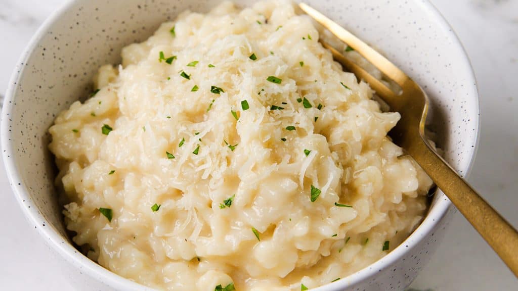A bowl of creamy Parmesan Risotto topped with grated cheese and chopped herbs, served with a gold fork.