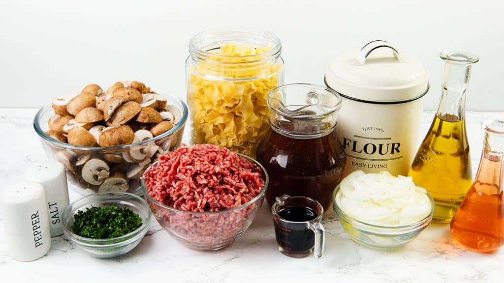Assorted ingredients for cooking a one pot meal, including ground beef, sliced mushrooms, egg noodles, chopped onions, parsley, flour, oil, broth, vinegar, salt, and pepper, arranged on a white countertop.