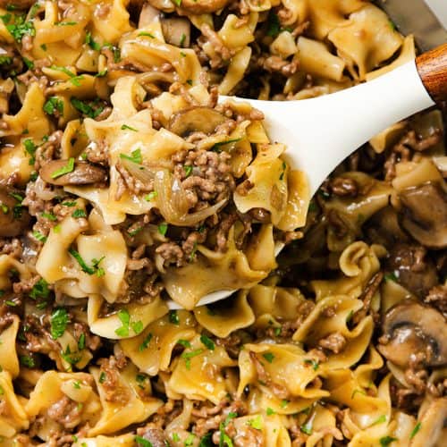 A one pot skillet brimming with egg noodles, ground beef, sliced mushrooms, and creamy sauce, garnished with chopped parsley. A white serving spoon lifts a hearty portion from the pan.