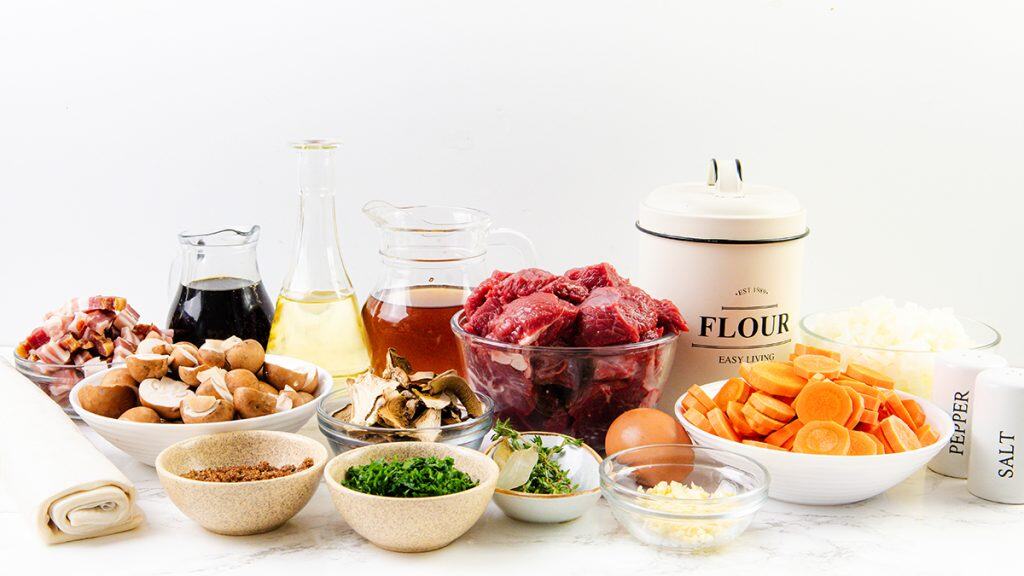 A variety of Beef and Guinness stew ingredients arranged on a white surface, including chopped beef, sliced carrots, onions, mushrooms, diced bacon, flour, eggs, parsley, garlic, broth, oil, soy sauce, salt and pepper.