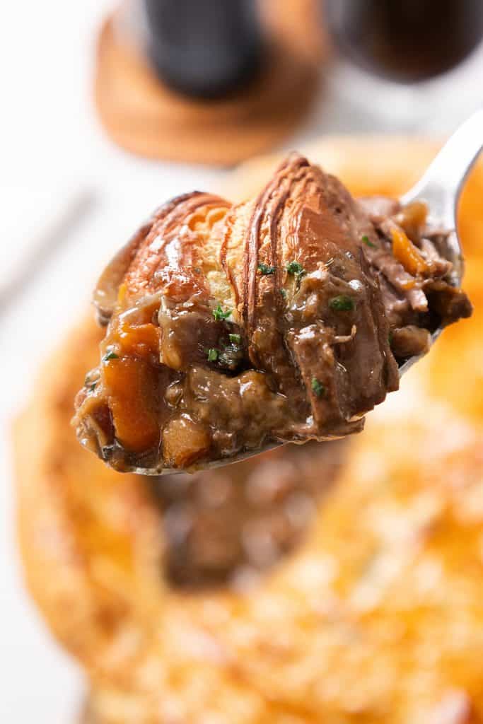 A close-up of a spoonful of savory beef and vegetable stew with carrots and herbs, featuring a golden-brown pastry crust in the background, suggesting this is a comforting beef and Guinness pie.