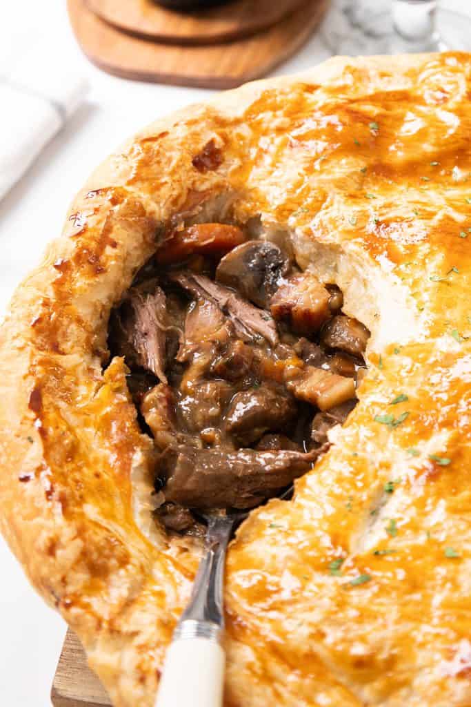 Close-up of a golden, flaky beef pie with a slice cut out, revealing a savory filling of tender beef, mushrooms, carrots, and rich Guinness gravy. A fork is inserted into the filling.