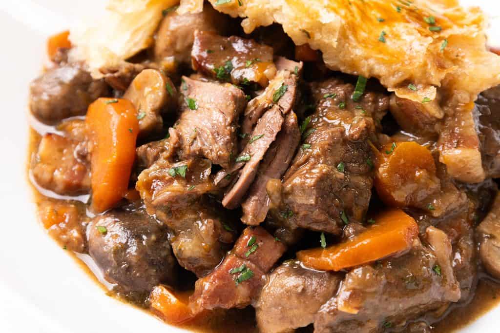 A close-up of a hearty Beef Pie with chunks of beef, carrots, mushrooms, and fresh herbs in a rich brown sauce, topped with a piece of golden, flaky pastry.