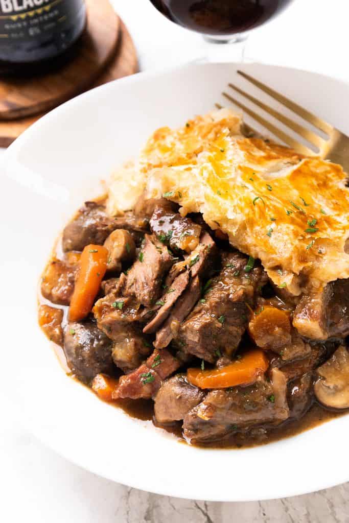 A white bowl filled with hearty Beef Pie stew, carrots, and mushrooms, topped with a golden, flaky pastry crust. A fork rests on the side, and a glass of red wine is visible in the background.