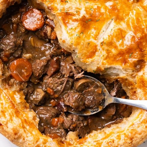 A golden, flaky Beef and Guinness Pie with a portion of crust removed reveals a rich beef and carrot filling. A spoon rests inside the pie, ready to serve.