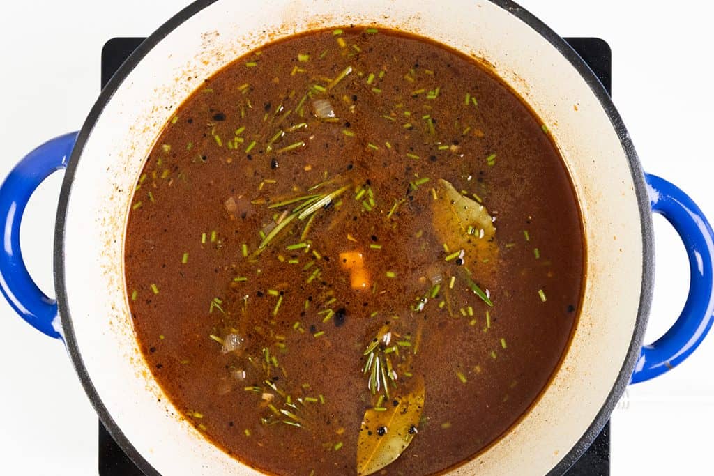A blue-handled pot filled with brown broth, braised lamb shanks, visible herbs, bay leaves, peppercorns, chopped onions, and carrots, seen from above on a white background.
