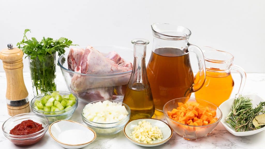 Assorted ingredients for a stew, including raw meat for braised lamb shanks, diced carrots, onions, celery, minced garlic, tomato paste, fresh herbs, olive oil, salt, pepper, and two pitchers of broth displayed on a white surface.
