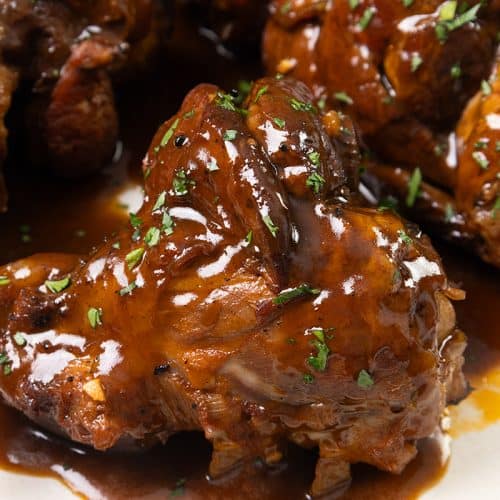 Close-up of braised lamb shanks simmered in sherry, covered in a rich, glossy brown sauce and garnished with chopped fresh herbs on a white plate.