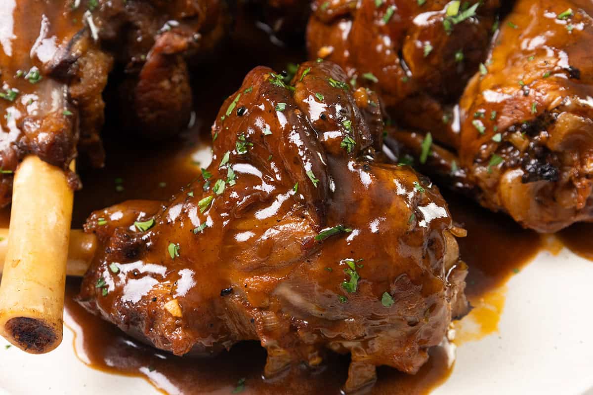 Close-up of braised lamb shanks simmered in sherry, covered in a rich, glossy brown sauce and garnished with chopped fresh herbs on a white plate.