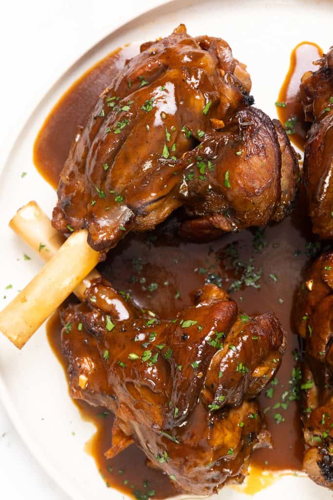 Close-up of braised lamb shanks covered in a rich sherry-infused brown sauce, garnished with chopped herbs and served on a white plate. The tender, glossy meat clings to the bones for an irresistible presentation.