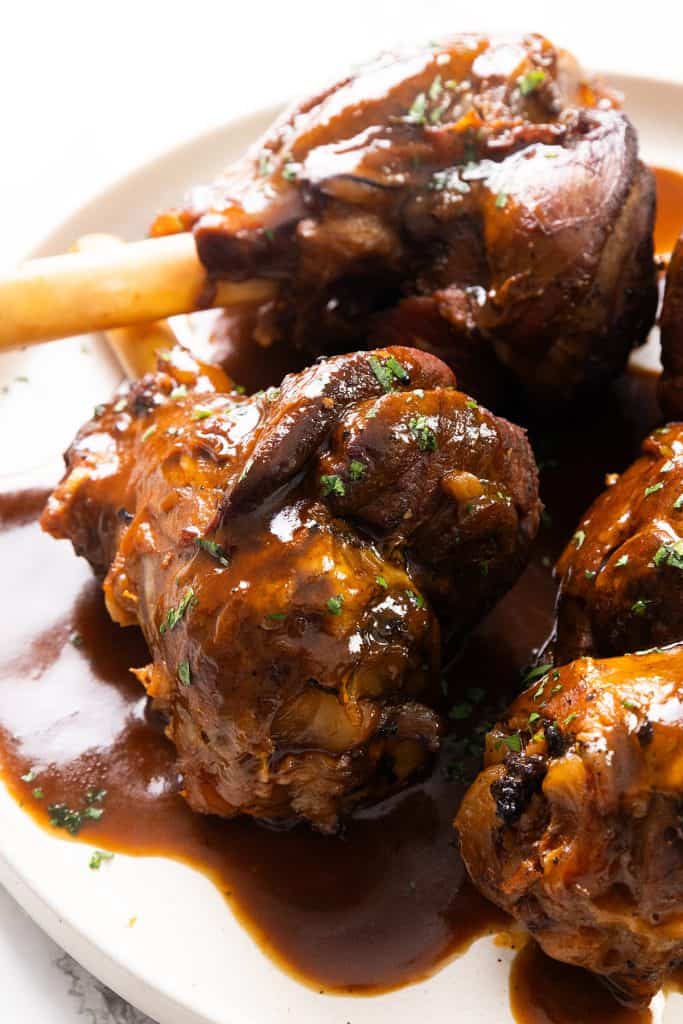 Three braised lamb shanks covered in a rich sherry-infused brown sauce are served on a white plate, garnished with finely chopped herbs. The meat appears tender and succulent.