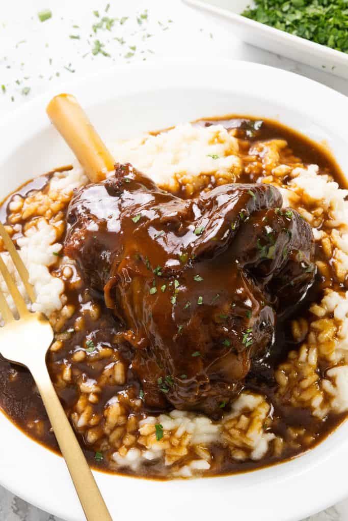 A braised lamb shank covered in rich brown gravy and a splash of sherry is served on creamy mashed potatoes in a white bowl, garnished with fresh herbs, with a gold fork on the side.
