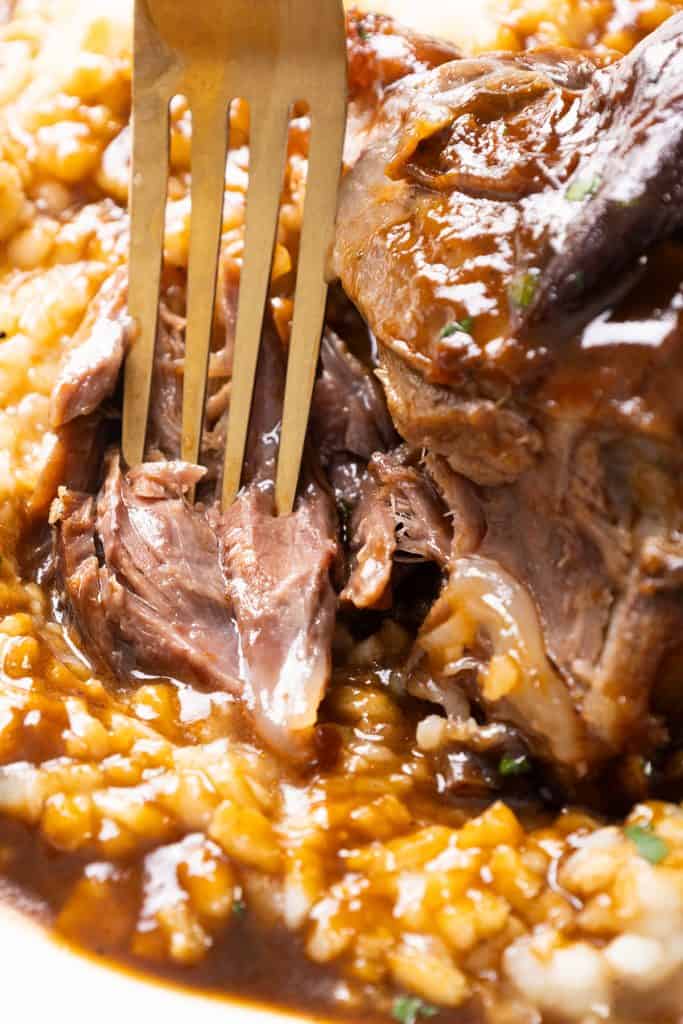 A close-up of a fork shredding tender braised lamb shanks on a bed of creamy risotto, all covered in rich brown gravy infused with herbs.