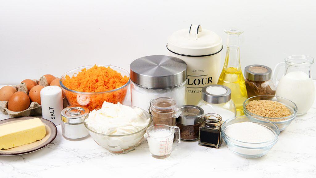 Various baking ingredients on a marble counter, including eggs, shredded carrots for carrot cake, butter, salt, cream cheese for cream cheese filling, oil, flour, sugars, milk, baking powder and soda, plus jars of spices.