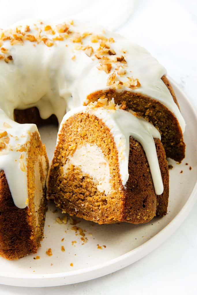 A carrot cake bundt with white icing and chopped nuts on top, sliced to reveal a creamy cream cheese filling inside, sits on a white plate.