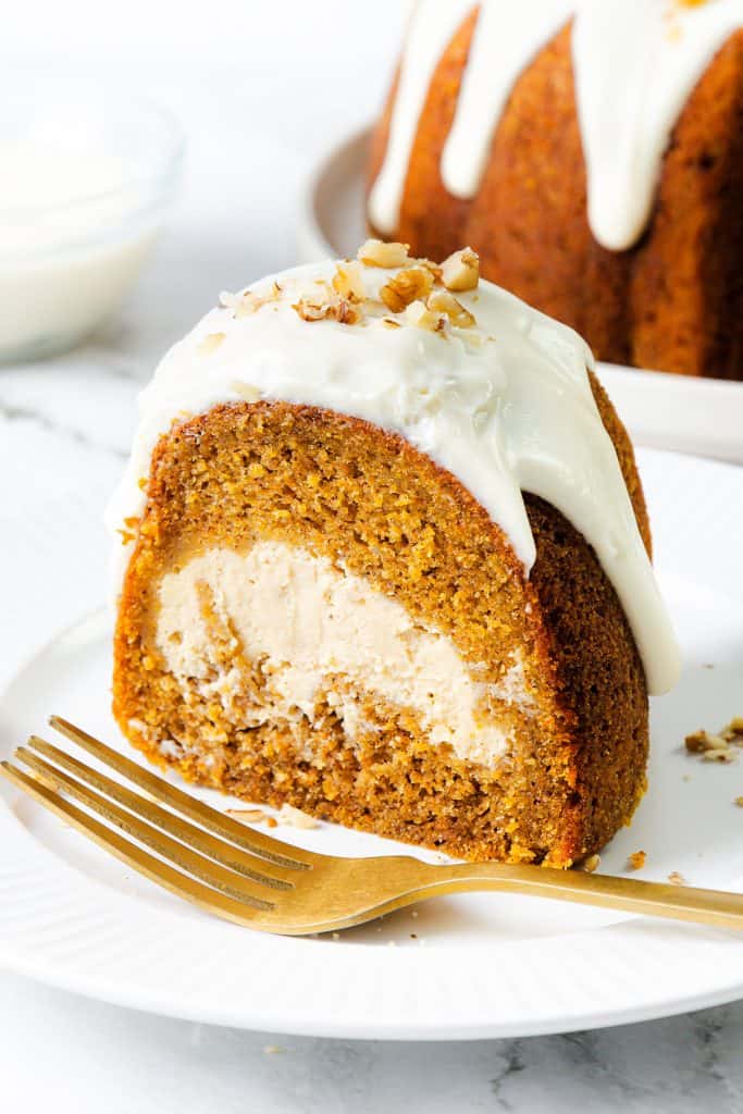 A slice of bundt cake with a cream cheese filling, topped with white icing and chopped nuts, sits on a white plate with a gold fork. The rest of the cake is visible in the background.