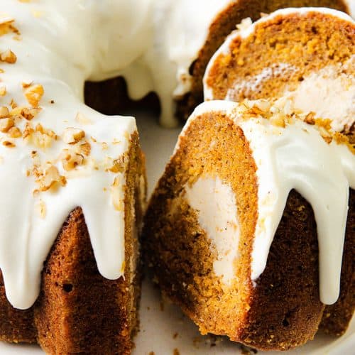 A pumpkin bundt cake with cream cheese filling, topped with white icing and sprinkled with chopped nuts, sits on a white plate. Two slices are cut to show the creamy center reminiscent of a classic carrot cake.