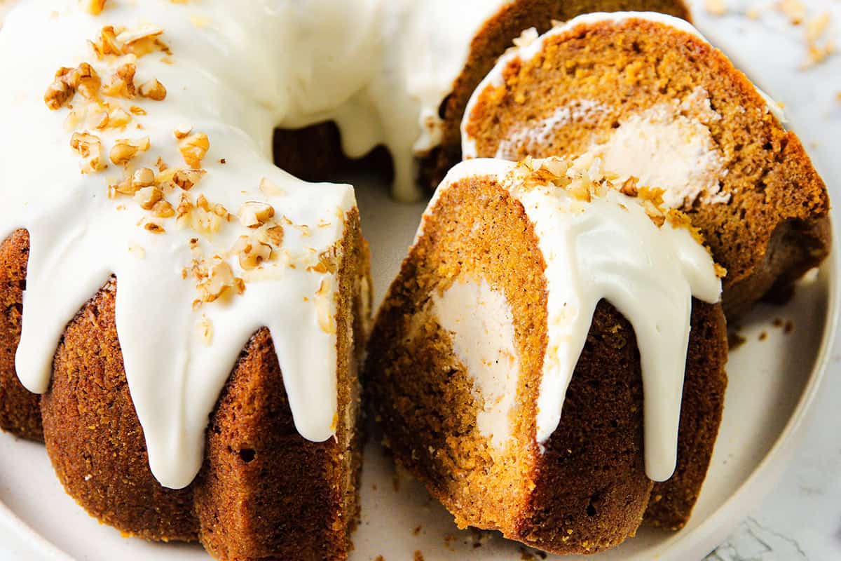 A pumpkin bundt cake with cream cheese filling, topped with white icing and sprinkled with chopped nuts, sits on a white plate. Two slices are cut to show the creamy center reminiscent of a classic carrot cake.